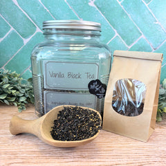 Vanilla black tea in a glass jar, wooden scoop, and paper bag on a wooden surface with a tiled wall background. Flavored black teas.