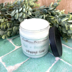 Whipped soap container with a black lid on a green tiled surface, surrounded by greenery.