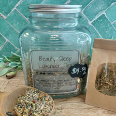 A clear glass jar labeled 'Beauty Sleep Lavender Tea' with a silver lid, shown on a wooden surface with a brown kraft paper pouch and a small wooden bowl of loose tea. A teal tiled wall is in the background.