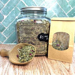 A clear glass jar labeled 'Blossoms of Health Tea' filled with loose herbal tea, shown on a wooden surface with a small wooden scoop in front and a kraft paper window box package to the right. A teal tiled wall is in the background.