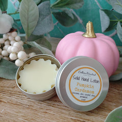 An open round metal tin of solid hand lotion and its lid, which displays the label 'Farmhouse Solid Hand Lotion Pumpkin Cardamom', styled on a wooden surface next to a pink decorative pumpkin and green leaves.