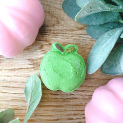 A small green apple-shaped item with a textured, dusty surface and a tiny brown stem detail, placed on a wooden surface with pink decorative objects and green leaves around it.