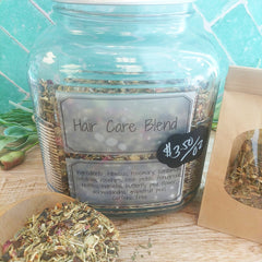 A clear glass jar labeled 'Hair Care Blend' filled with dried herbs and flowers, shown on a wooden surface with a kraft paper pouch and loose blend in front. The label includes a price tag graphic.