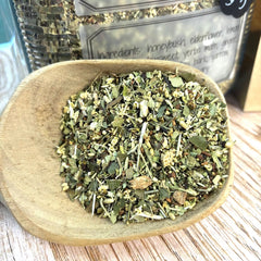 A close-up of loose herbal tea or dried botanicals in a small wooden spoon, with a branded jar blurred in the background on a light wood surface.
