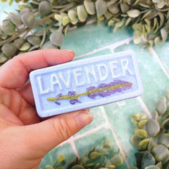 Embroidered patch with 'LAVENDER' text and lavender design held by a hand against a green leafy background.