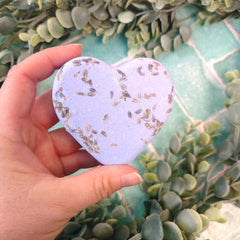 Heart-shaped bath bomb with decorative elements held in a hand against a green leafy background