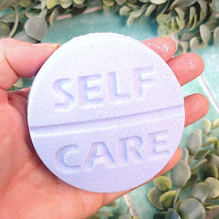 Hand holding a lavender 'SELF CARE' pill-shaped object with greenery in the background