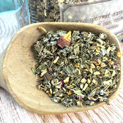 A close-up of loose-leaf herbal tea mixed in a light wood spoon, with dried botanical pieces visible against a rustic tabletop background.