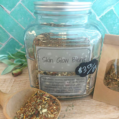 A clear glass jar labeled 'Skin Glow Blend' filled with loose herbal tea mix, shown on a wooden surface with a small wooden spoon of the blend in front and a kraft paper pouch beside it. A turquoise tiled wall is in the background.
