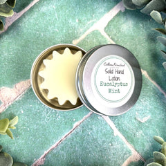 Solid hand lotion in a metal tin with a label on a green leafy background. Calhoun Farmstead DuBois Pa. Herb store.
