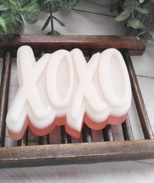 Goats milk and glycerin soap. Shaped like XOXO hugs and kisses. Soap is white on top with red bottom. Soap is sitting on a wooden soap dish.